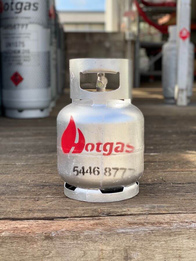 A small silver gas cylinder labeled "Hotgas" with a red flame logo stands on a wooden pallet. Other similar gas cylinders are blurred in the background. The number "5446 877" is printed on the cylinder.