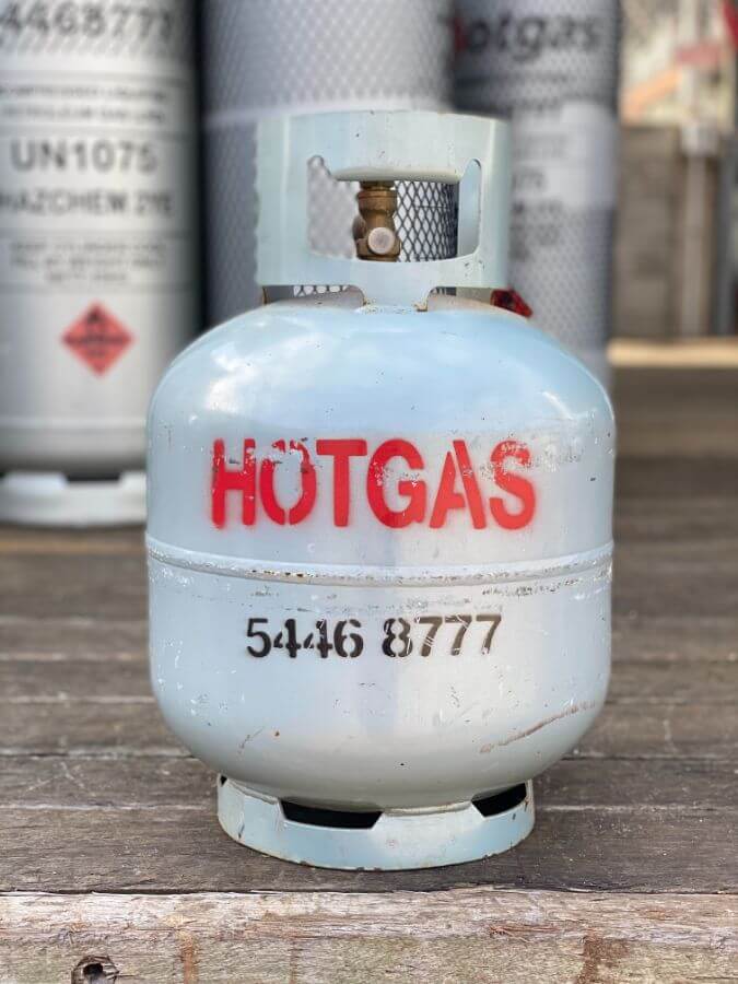 A metal gas cylinder with "HOTGAS" stenciled in red letters and the phone number "5446 8777" written below it. Several similar gas cylinders are seen in the background. The cylinder is placed on a wooden surface.