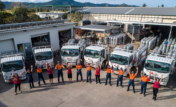 Hotgas Team A group of uniformed people stands in front of several trucks with raised hands, forming an arc. The trucks are parked in an industrial area with storage tanks and warehouse buildings in the background. The landscape features trees and hills.