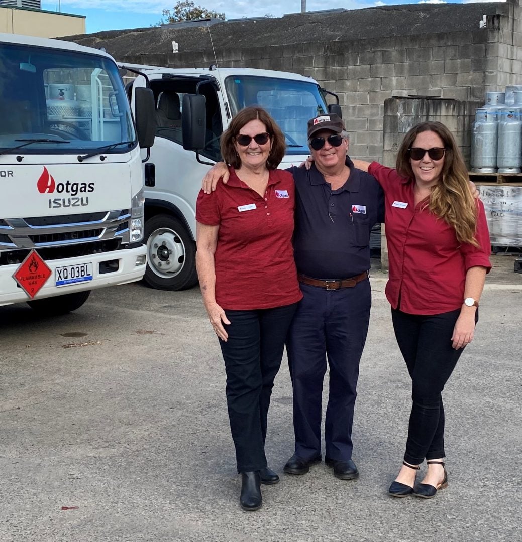 Three employees stand smiling in front of an Otgas Isuzu truck. The two women are wearing red polo shirts and black pants, while the man in the middle wears a dark uniform. They are posing in an industrial area with gas cylinders in the background—your LPG specialists celebrate 30 years!